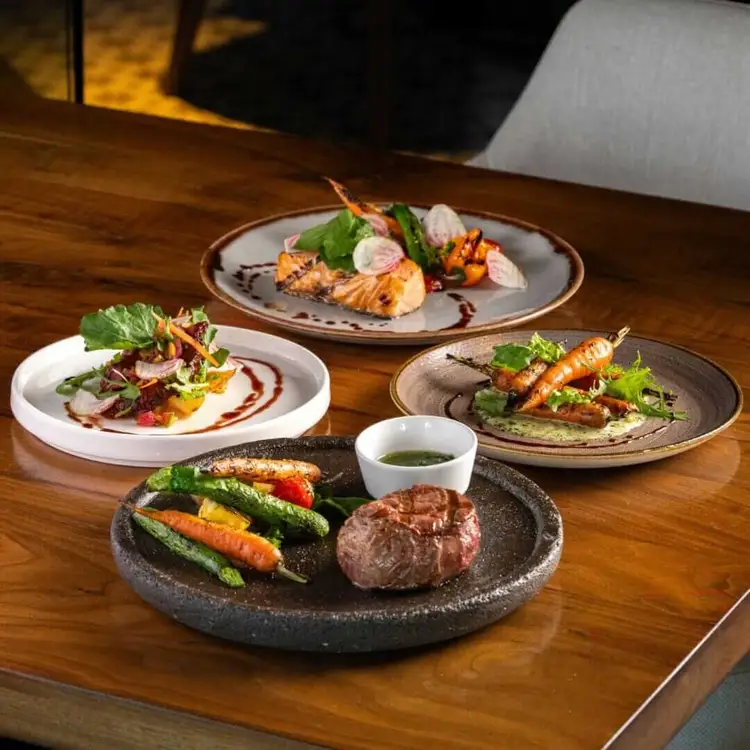 Four plated dishes arranged on a wooden table featuring grilled proteins, roasted vegetables, fresh herbs, and sauce garnishes Four plated dishes arranged on a wooden table featuring grilled proteins, roasted vegetables, fresh herbs, and sauce garnishes