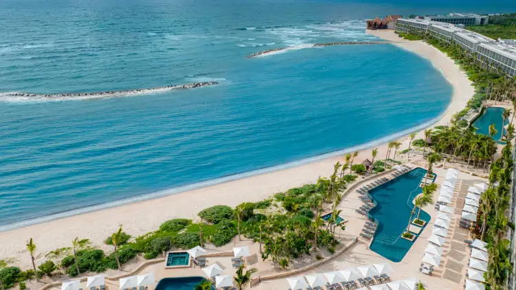 Aerial view of a resort with sandy beach, turquoise water, swimming pools, palm trees, and multi-story hotel buildings along the shoreline hitlon-tulum-lsyn