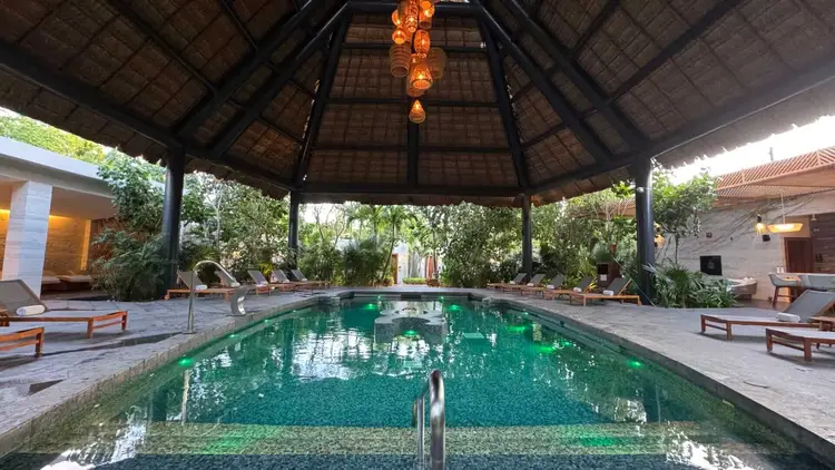 A rectangular pool with turquoise water sits under a thatched roof pavilion surrounded by lounge chairs and tropical vegetation A rectangular pool with turquoise water sits under a thatched roof pavilion surrounded by lounge chairs and tropical vegetation