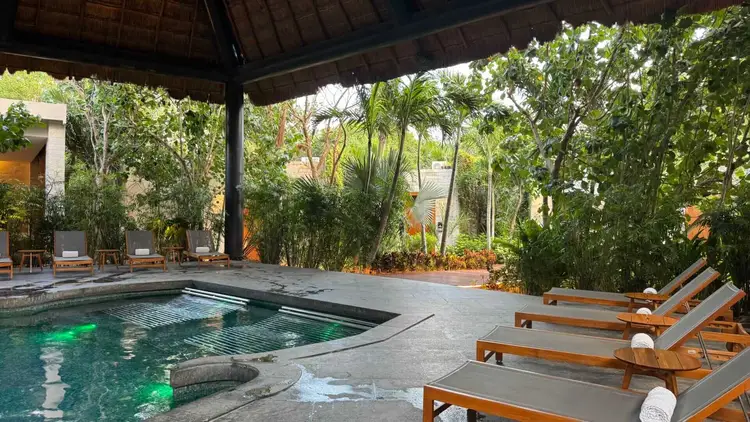 Wooden lounge chairs line the deck beside an illuminated pool under a covered pavilion surrounded by tropical plants and trees Wooden lounge chairs line the deck beside an illuminated pool under a covered pavilion surrounded by tropical plants and trees