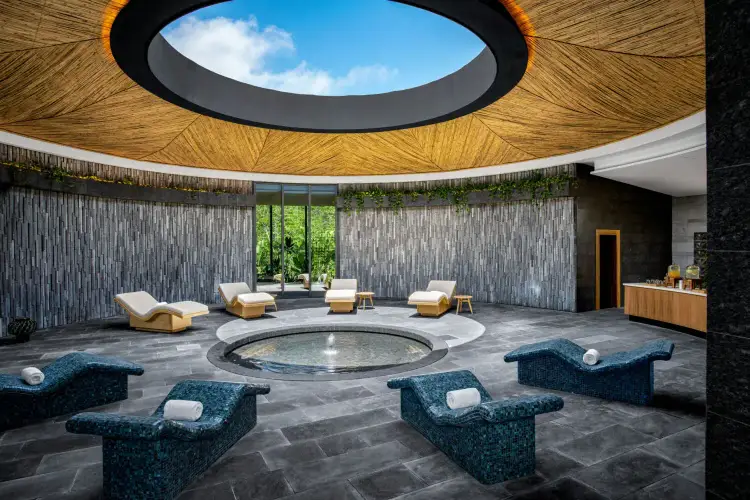 Modern spa interior with a circular pool in the center, blue sculptural seating, yellow loungers, and an oculus opening to the sky in the wooden ceiling conrad-tulum3-1
