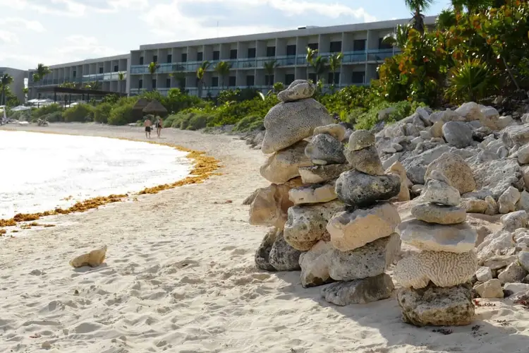 Large stacked rocks line a sandy beach with a multi-story waterfront building and vegetation in the background Large stacked rocks line a sandy beach with a multi-story waterfront building and vegetation in the background