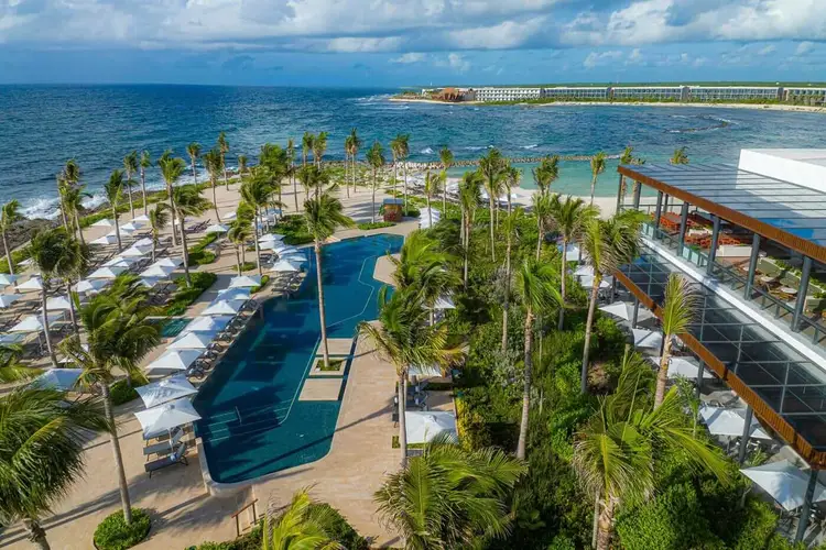 Aerial view of a beachfront resort with a blue pool surrounded by palm trees, white lounge chairs, sandy beach, and ocean beyond riv-hilton-tulum-riviera-maya-aerial-03