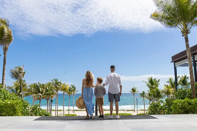Family of three stands on a beachfront promenade looking toward the ocean, with palm trees and sandy beach visible in the background Family of three stands on a beachfront promenade looking toward the ocean, with palm trees and sandy beach visible in the background