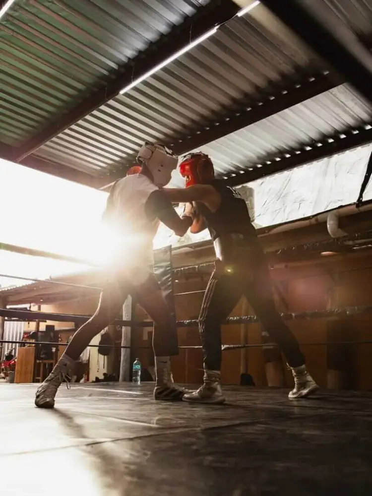 Two boxers spar in a ring inside a covered training facility with corrugated metal roof panels and bright sunlight streaming in
