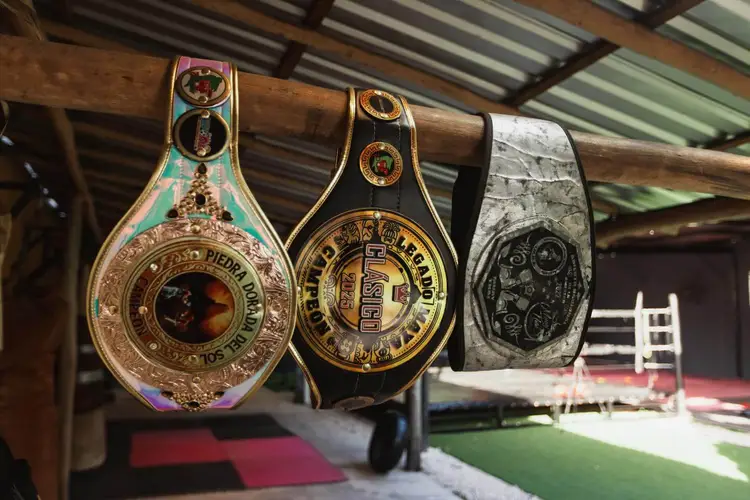 Three ornate championship boxing belts hang from a wooden beam inside a venue with a boxing ring visible in the background