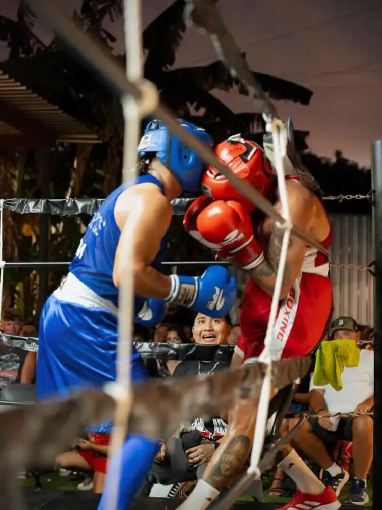 Two boxers in blue and red gear exchange punches in a boxing ring while spectators watch from behind the ropes at night