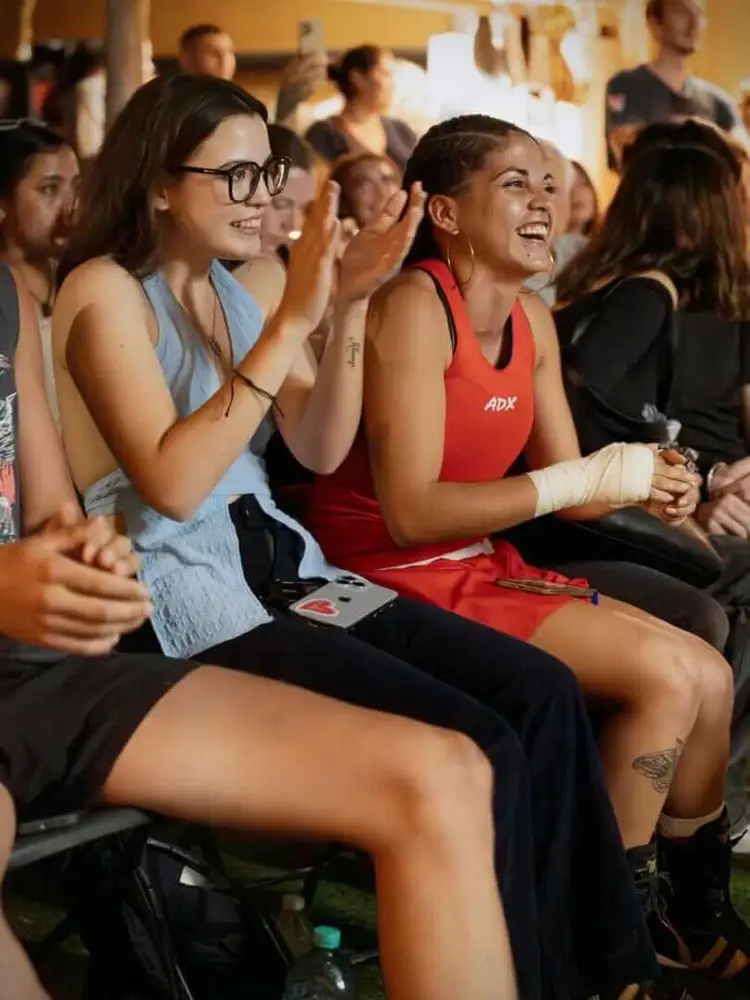 A group of people watch boxing matches indoors, smiling and clapping as spectators at an event held in a yellow-walled venue