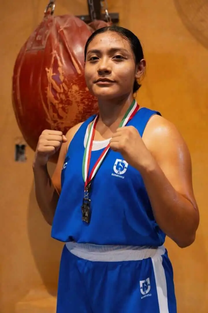 Boxer wearing blue athletic gear and a medal stands holding a heavy bag against an orange wall