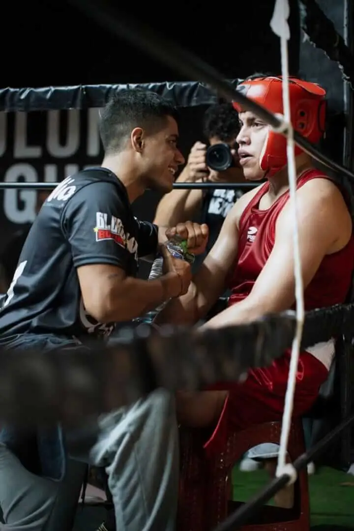 Boxer in red headgear speaks with coach in black shirt at boxing event ringside