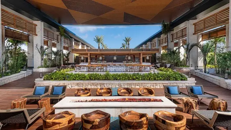 A resort courtyard with a sunken fire pit surrounded by wooden seating, lounge chairs, and potted plants, with an open-air bar and palm trees visible in the bac 482321039_675624934975684_5862340575962945904_n