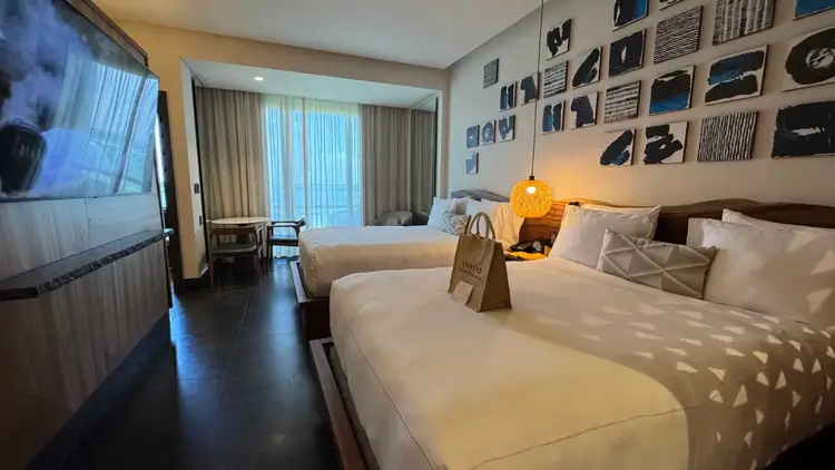 Hotel room with two beds, a television, and framed artwork on the walls, opening onto a balcony with curtained doors Hotel room with two beds, a television, and framed artwork on the walls, opening onto a balcony with curtained doors