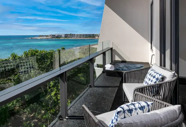 Waterfront balcony with lounge chairs overlooking a turquoise beach and resort buildings under blue sky HABITACIONES-FS