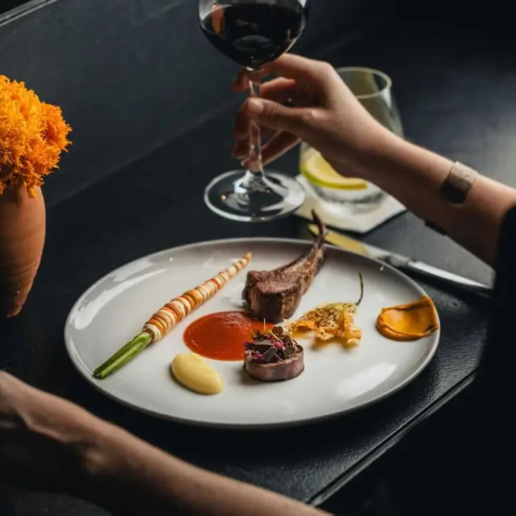 A person holds a glass of red wine above a plated gourmet meal with meat, sauce, and garnishes on a dark table with yellow flowers nearby 465768301_18004093313659103_3564317435233917773_n