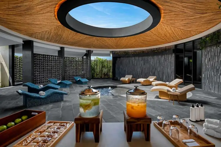 Modern spa facility with circular skylight, lounging areas with blue cushioned seating, beverage stations with infused drinks, and towel service around a slate Conrad-Spa-Relaxation-Area-2