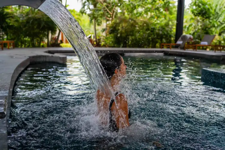 A person sits in a pool as water pours over their head from a wooden trough in a tropical resort setting Mayo-2024_Conrad-255