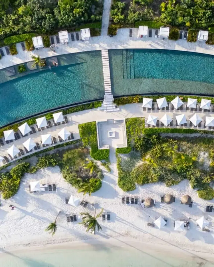 Aerial view of a resort with curved swimming pools, white loungers on sandy areas, palm trees, and a beachfront with turquoise water Aerial view of a resort with curved swimming pools, white loungers on sandy areas, palm trees, and a beachfront with turquoise water