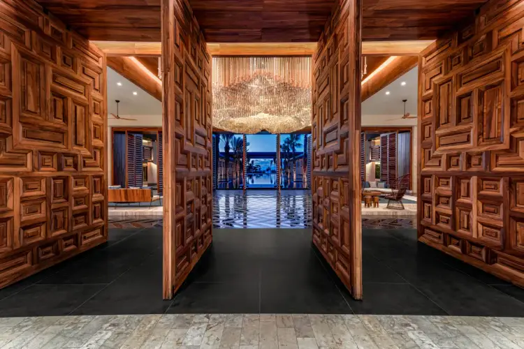 Ornate wooden doors with geometric patterns frame an entrance to a modern luxury interior with water views visible through floor-to-ceiling windows at dusk Conrad Tulum_©Victor Elias_Lobby (3) (1)
