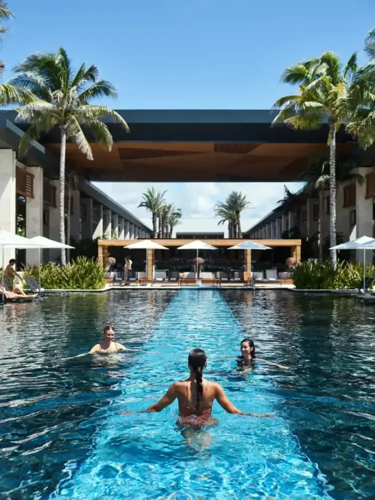 Guests swim in a long rectangular pool lined with palm trees at a resort with modern architecture and covered lounge areas MAIN-POOL_0844-v2