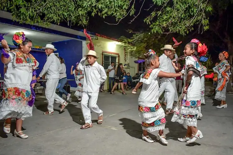 People in colorful traditional clothing and straw hats dance together in a street at night outside a blue building