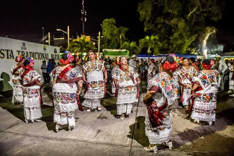 People wearing white embroidered traditional clothing with red floral patterns gather on a nighttime street at an outdoor event