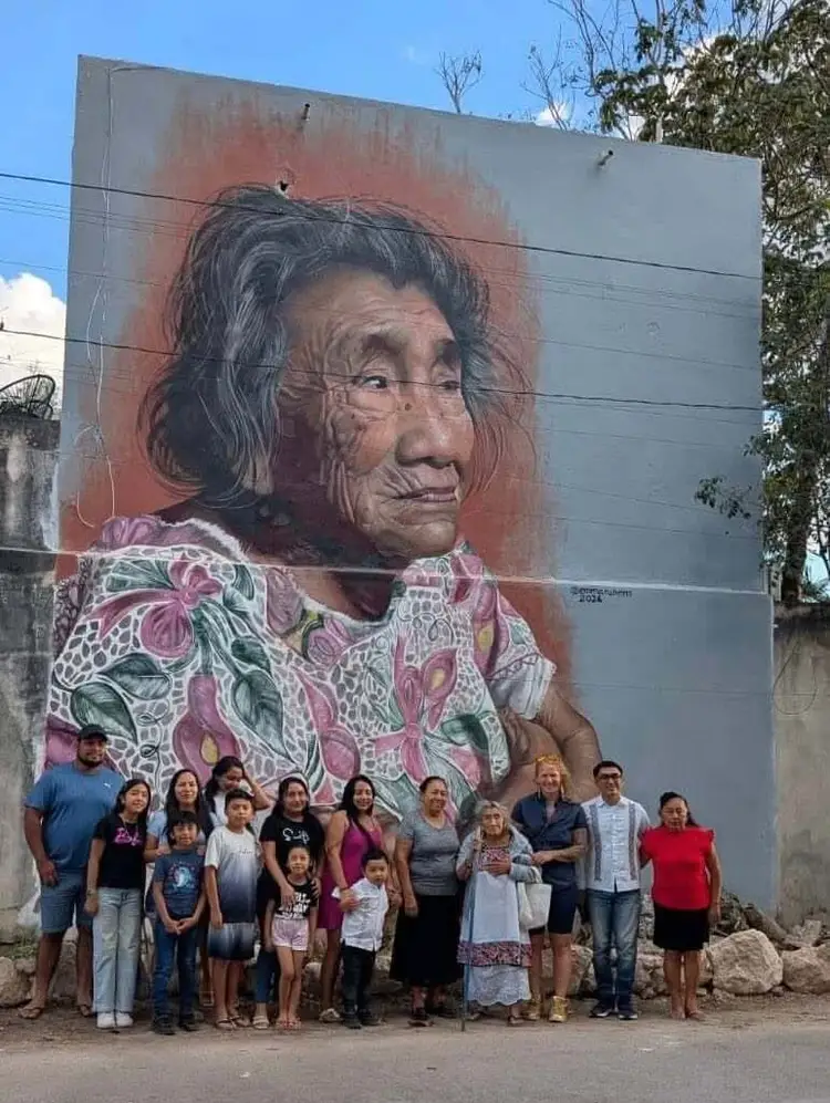 A group of people of various ages stand together in front of a large mural depicting an elderly woman wearing a floral patterned shirt 646611304_1351794706995014_8740606993614490778_n