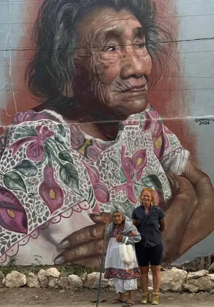Two women stand in front of a large mural depicting an elderly woman wearing glasses and a colorful patterned garment Two women stand in front of a large mural depicting an elderly woman wearing glasses and a colorful patterned garment
