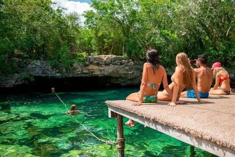 Group of people sitting and swimming at a cenote with turquoise water surrounded by limestone cliffs and tropical vegetation cenote azul