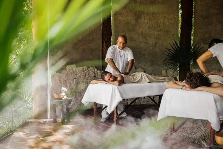 Massage therapists work with clients in a spa treatment room with stone walls and hanging plants