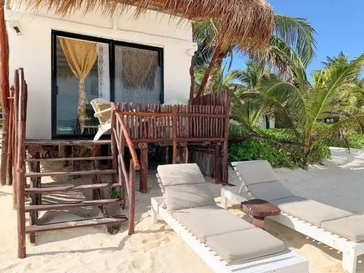 Beach bungalow with thatched roof and wooden deck overlooking lounge chairs on sand with palm trees nearby