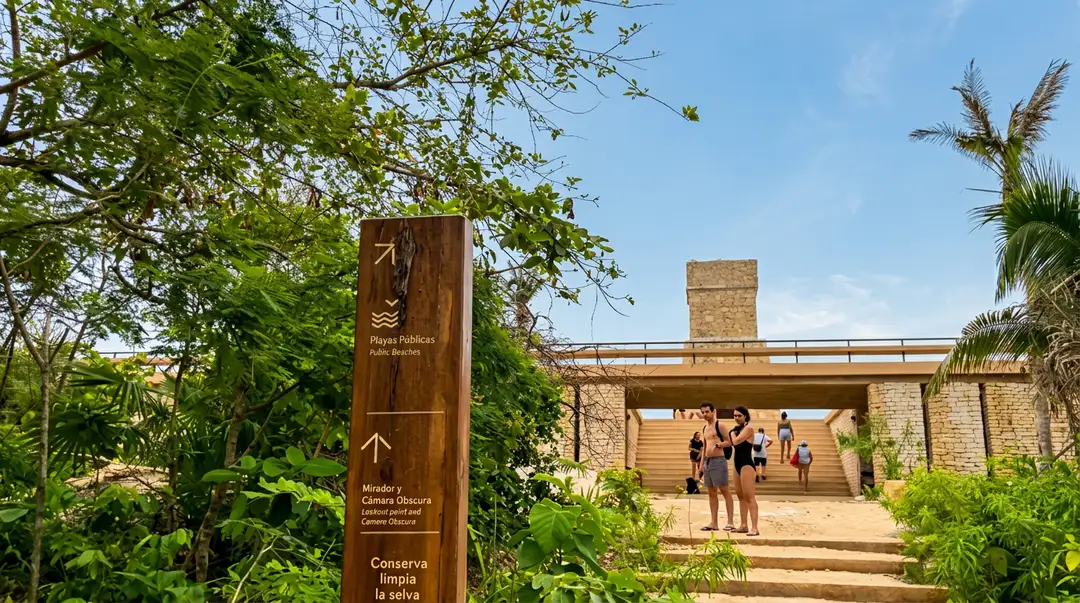 A wooden directional sign stands among tropical vegetation near a modern brick building where visitors gather under a covered structure