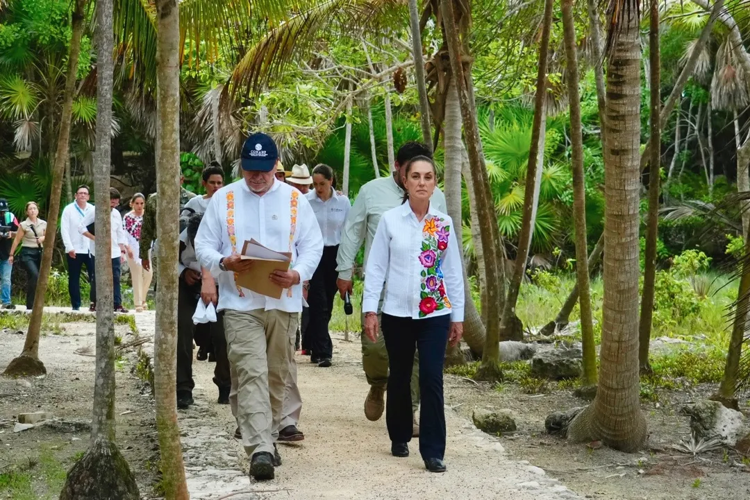 A man in a cap carrying a wooden box and a woman in a floral-embroidered shirt walk along a sandy path through a dense palm forest with other visitors behind th