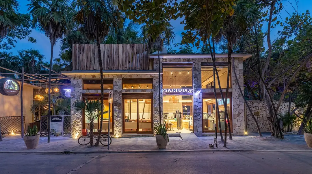 Starbucks closes its Tulum hotel zone store after 11 years Illuminated Starbucks storefront with stone and wood facade surrounded by palm trees and tropical vegetation at dusk