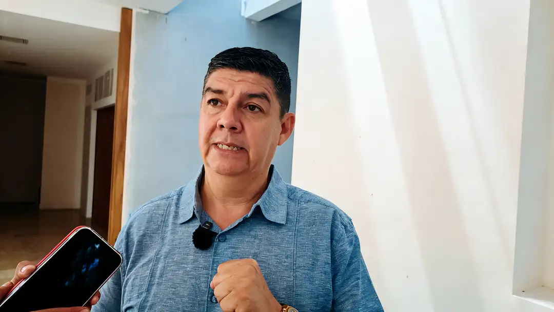 Nuevas colonias, nuevas calles y ninguna señal que las regule Man in blue shirt holds a phone while speaking indoors, with a hallway and blue wall visible behind him