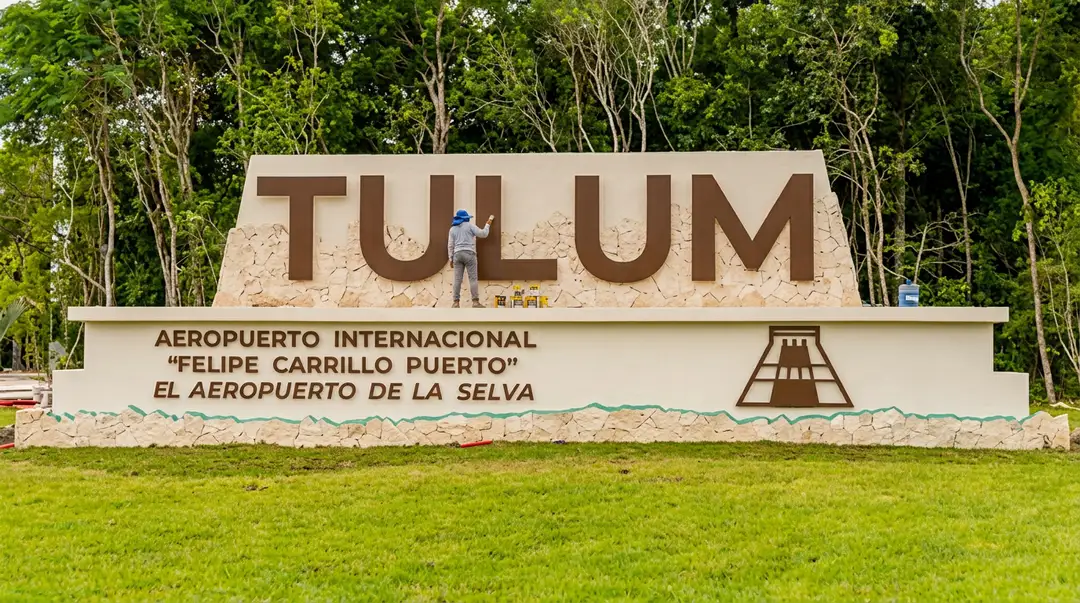 Tulum paga la factura de imagen de un aeropuerto en otro municipio Large sign reading "TULUM" with text referencing an international airport below it, set against a forested backdrop with grass in the foreground