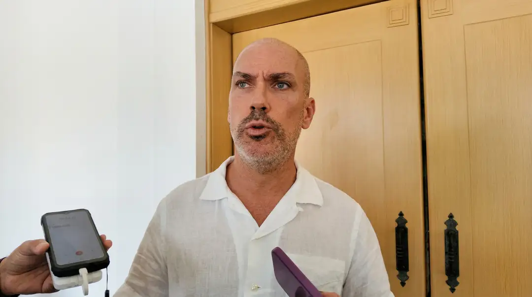 Tulum se hunde y el estado no hace nada Man in white shirt holds a smartphone while speaking indoors in front of wooden doors