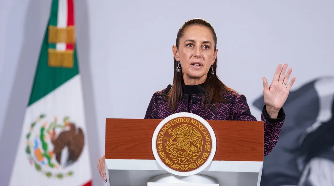 Woman speaking at a podium bearing an official seal, with a Mexican flag visible in the background