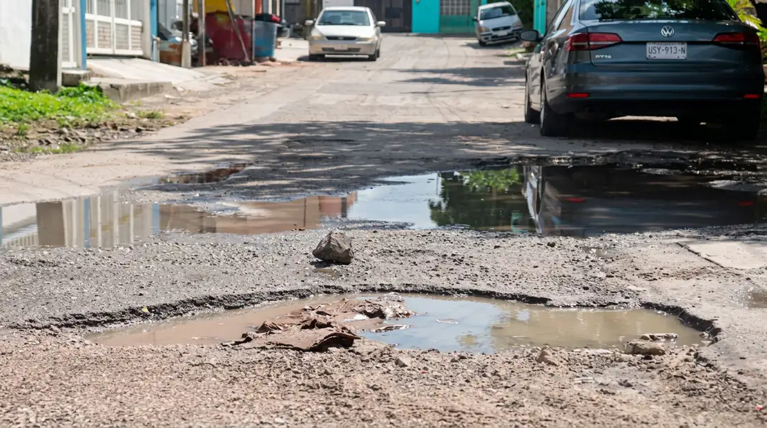 A large pothole filled with water sits in a deteriorated street lined with parked cars and residential buildings