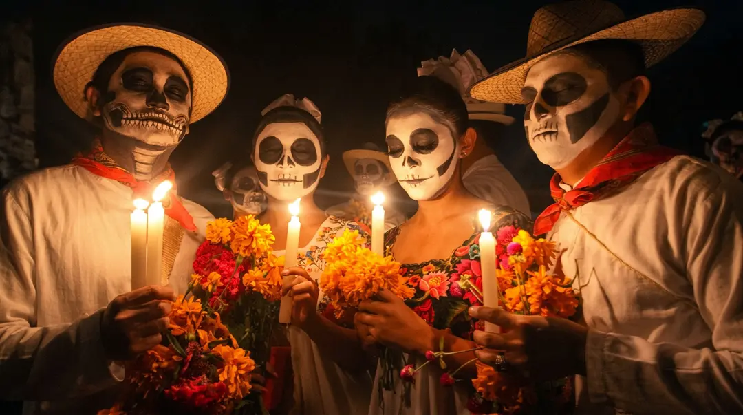 Four people wearing skull face paint and wide-brimmed hats hold candles and orange flowers while posing together at night