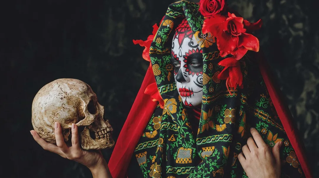 Person wearing skull face makeup and ornate green embroidered clothing with red fabric holds a human skull prop against a dark background