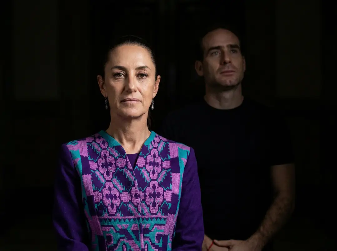 Woman wearing a purple and pink patterned vest stands facing forward against a dark background, with a man in a black shirt visible behind her