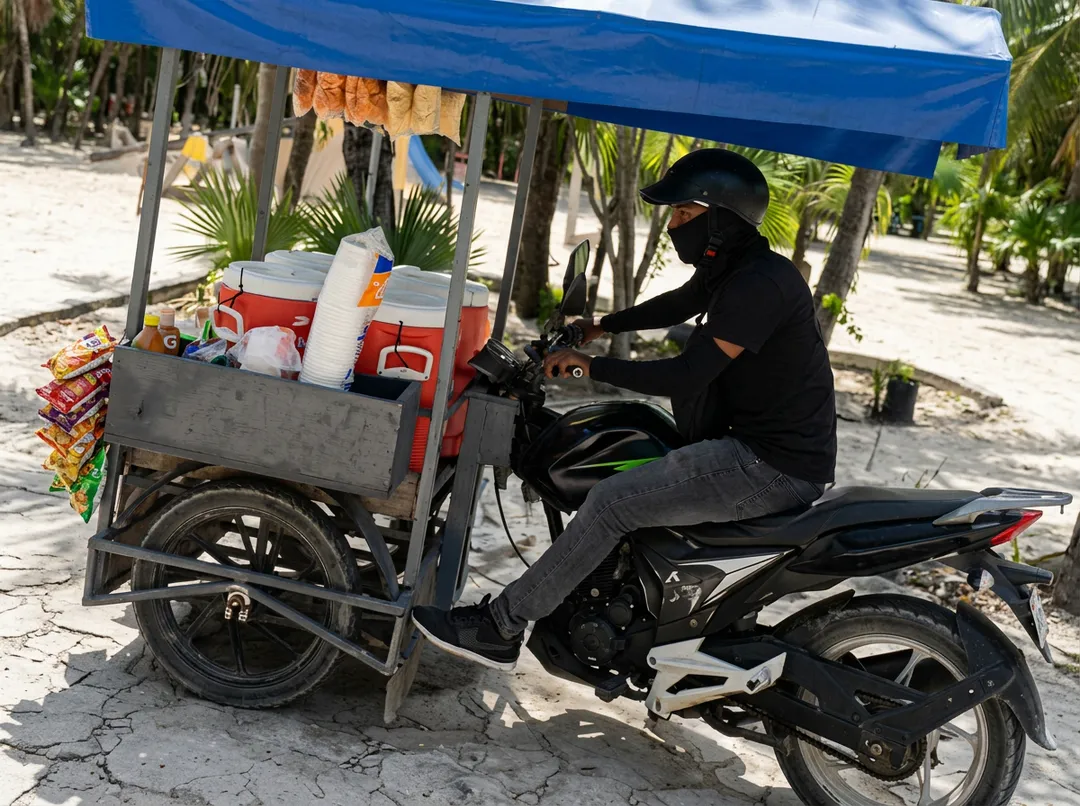 A vendor wearing a black helmet and mask sits on a motorcycle next to a food cart with a blue umbrella, stocked with coolers and packaged snacks under palm tree