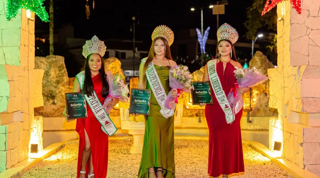 Three women wearing formal gowns, decorative headpieces, and sashes hold flowers and certificates while posing in an illuminated archway at night