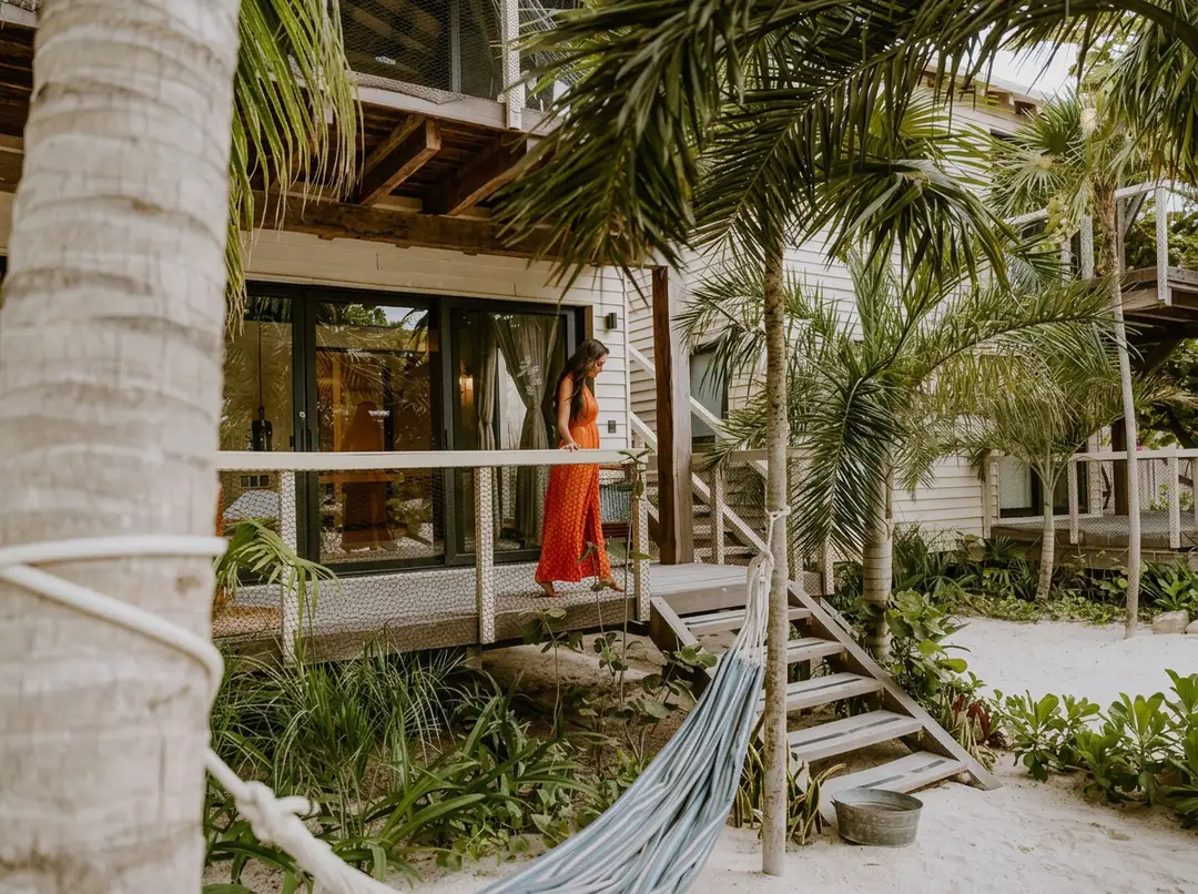 Woman in orange dress stands on a beachfront home's screened porch surrounded by palm trees and tropical vegetation