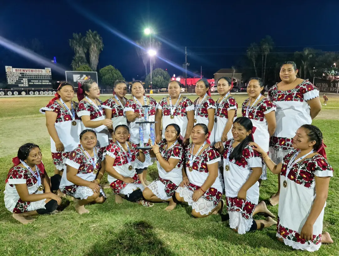 A women's softball team in matching red and white uniforms poses together on a grass field at night holding a trophy