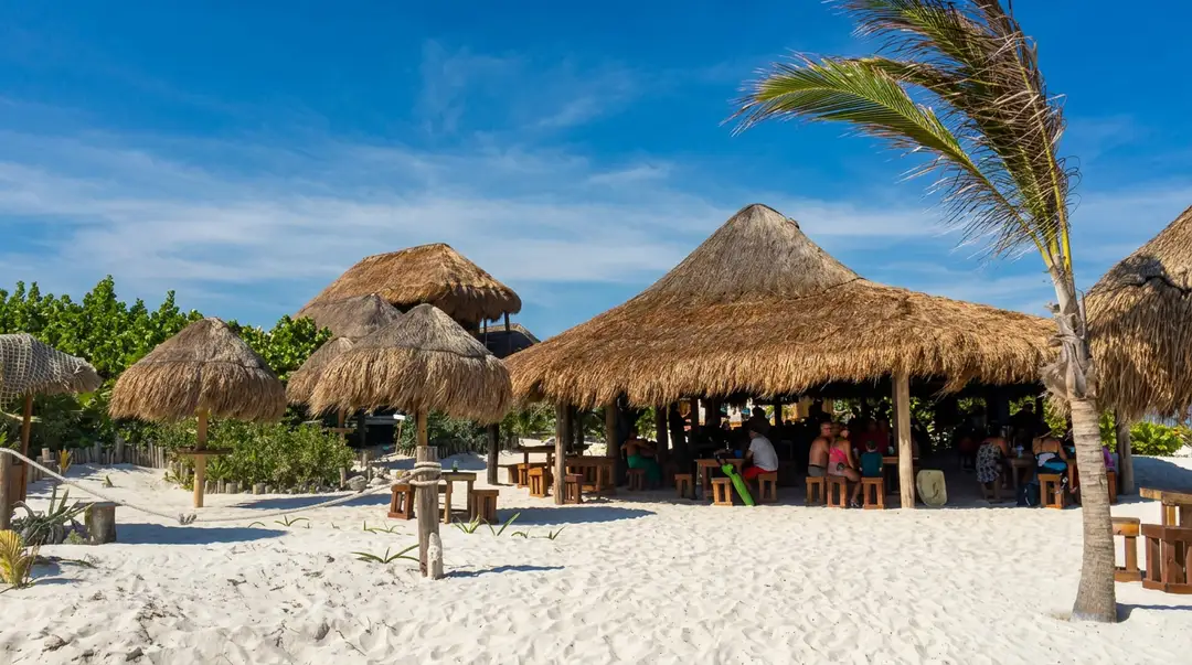 Thatched-roof beach structures and palm trees sit on white sand with visitors gathered near the buildings under a clear blue sky