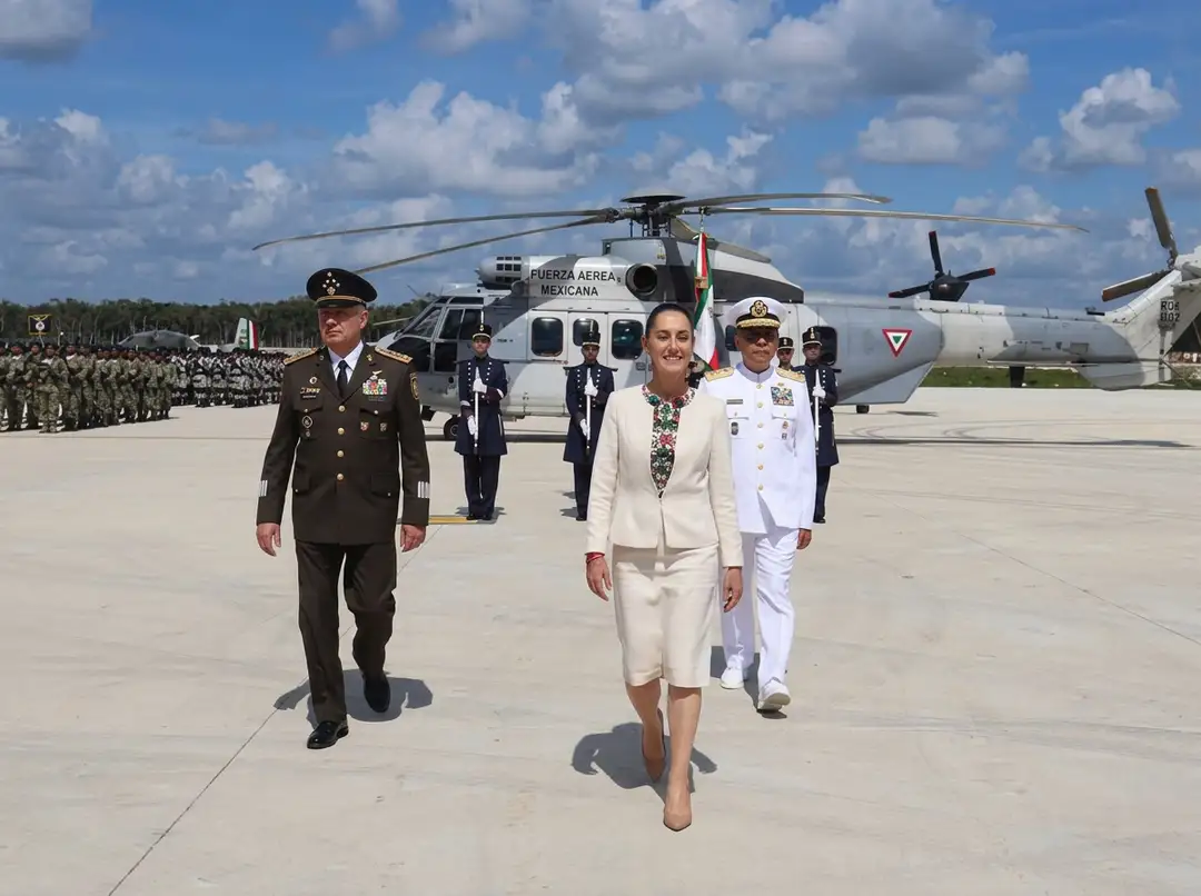 Military and civilian officials walk on a tarmac in front of a helicopter with uniformed personnel and troops in the background