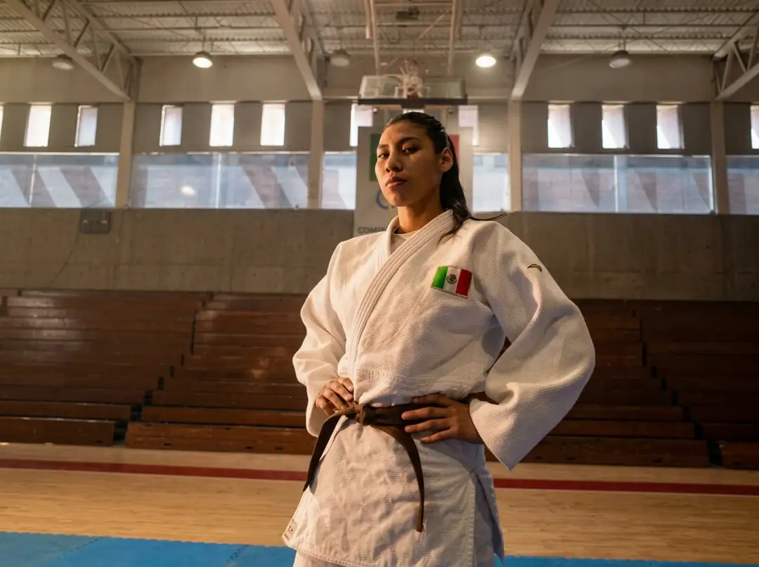 Judo athlete in white gi and black belt stands on a mat inside a gymnasium with a Mexican flag patch on her uniform