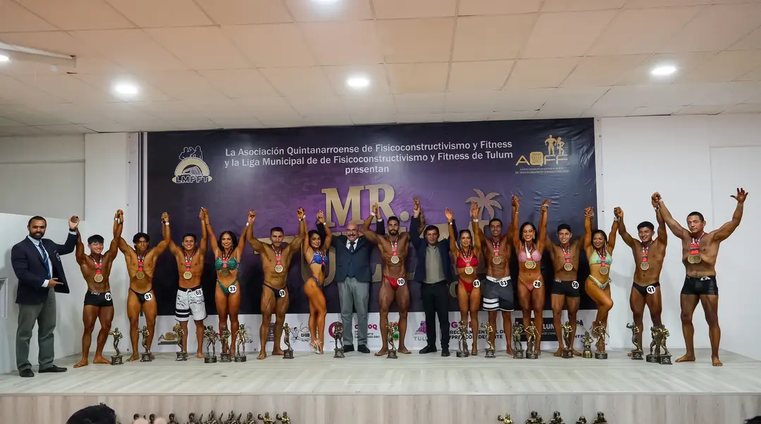 Bodybuilders in competition attire and medal winners stand on stage with their arms raised at a fitness competition event