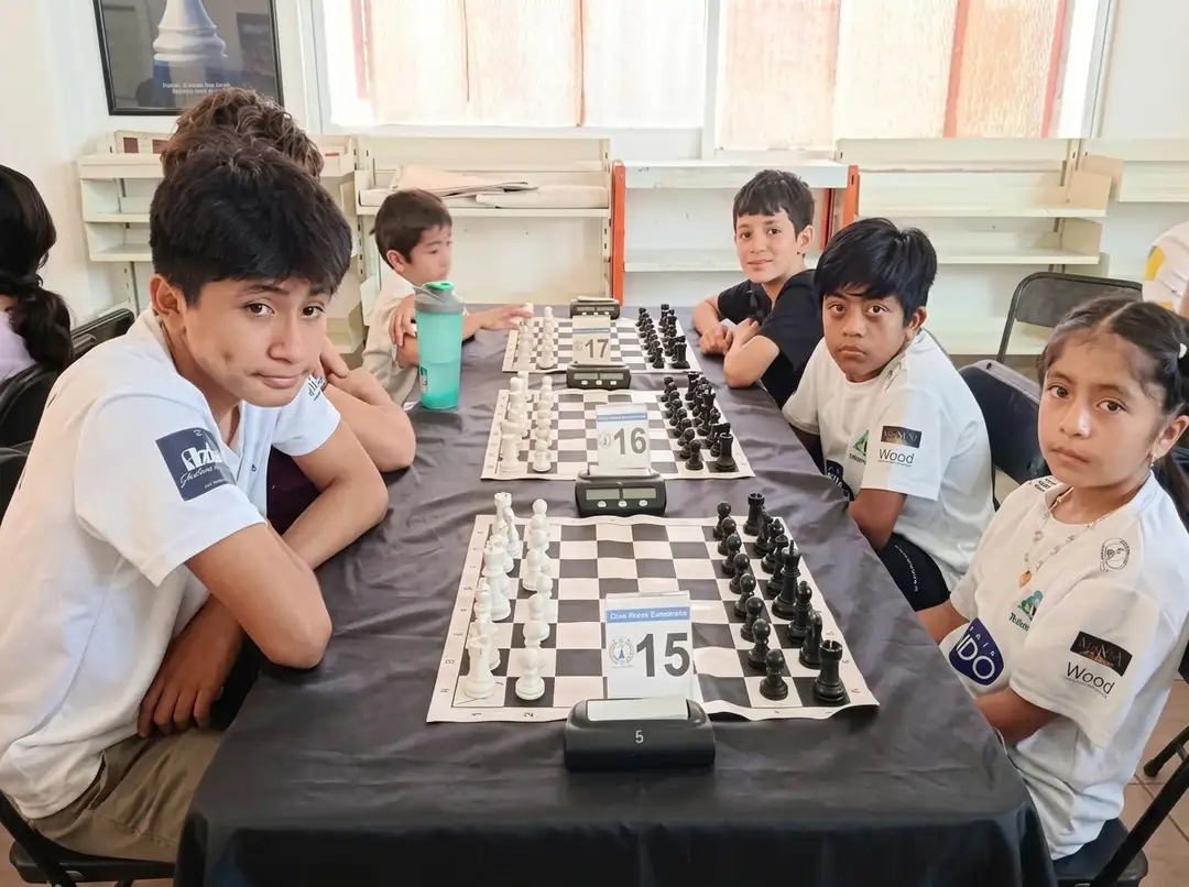 Young chess players sit at a table with multiple chess boards during a tournament competition indoors